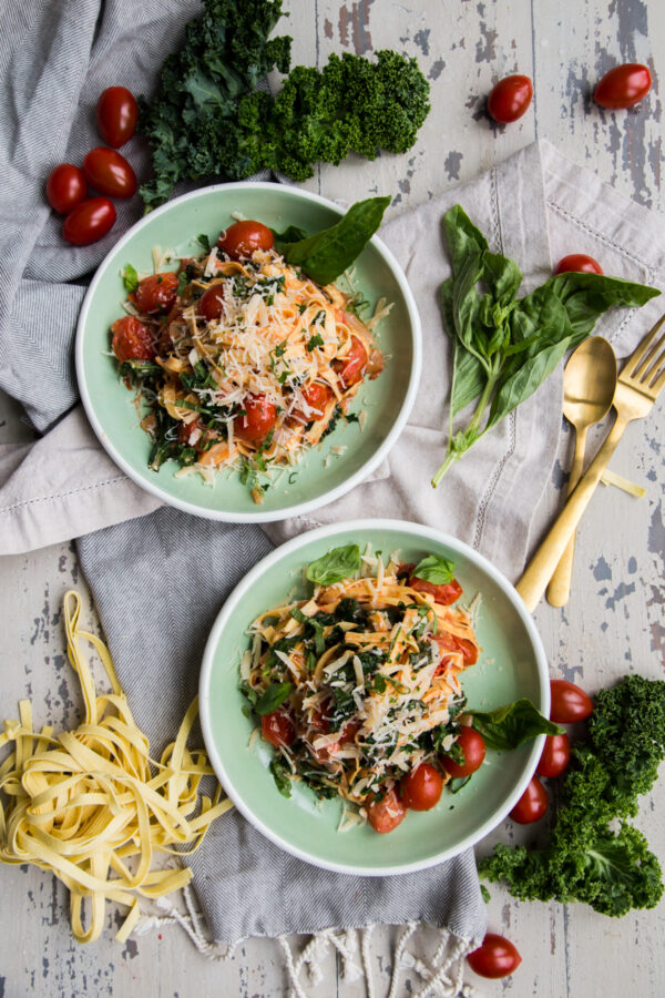 Rustic Tomato Kale Alfredo Pasta overhead on green plates surrounded by fresh pasta gold utensils and fresh kale