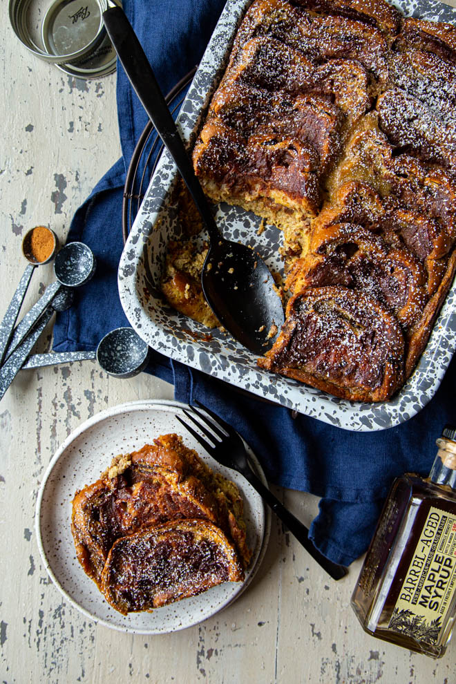baked french toast in a gray and white speckled dish with dish and bottle of maple syrup, shot overhead