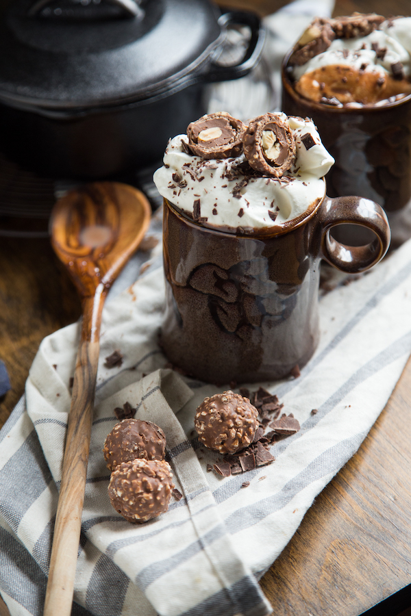 Nutella hot cocoa in a mug with whipped cream, with chocolate surrounding it and a cast iron pot in the background