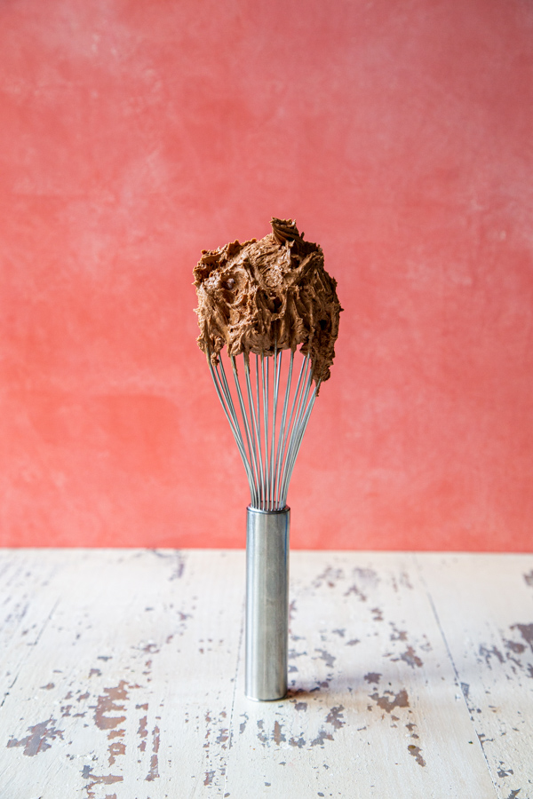 How to make chocolate buttercream tutorial, with a whisk of chocolate butter cream on a pink background and a white foreground