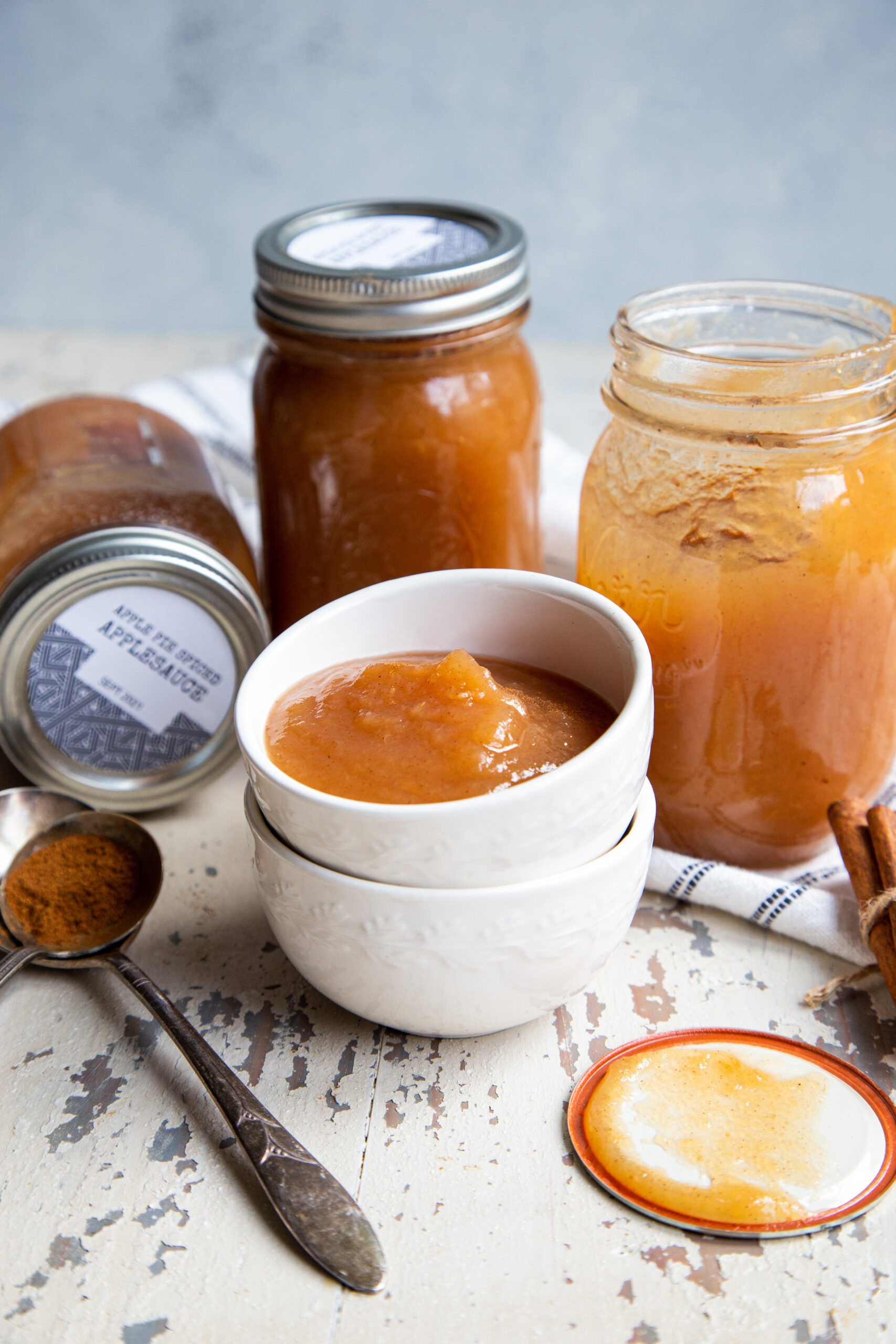 Three glass mason jars sitting behind two nested white ceramic bowls filled with applesauce and spoon holding ground cinnamon