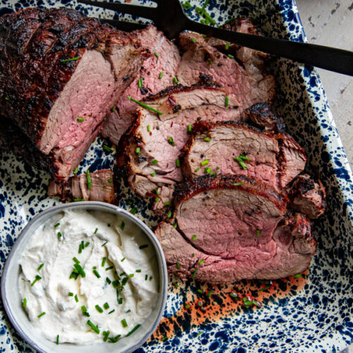 Sliced grilled beef tenderloin with serving fork and bowl of horseradish chive sauce