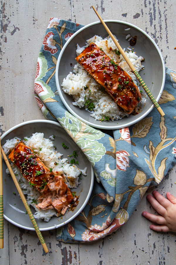 Overhead view of two plates of teriyaki glazed herb salmon with rice, with toddler hand in foreground